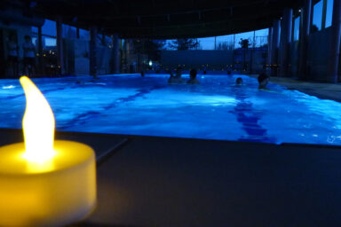 Bougie au premier plan et piscine avec des personnes en train de se relaxer dans la pénombre.